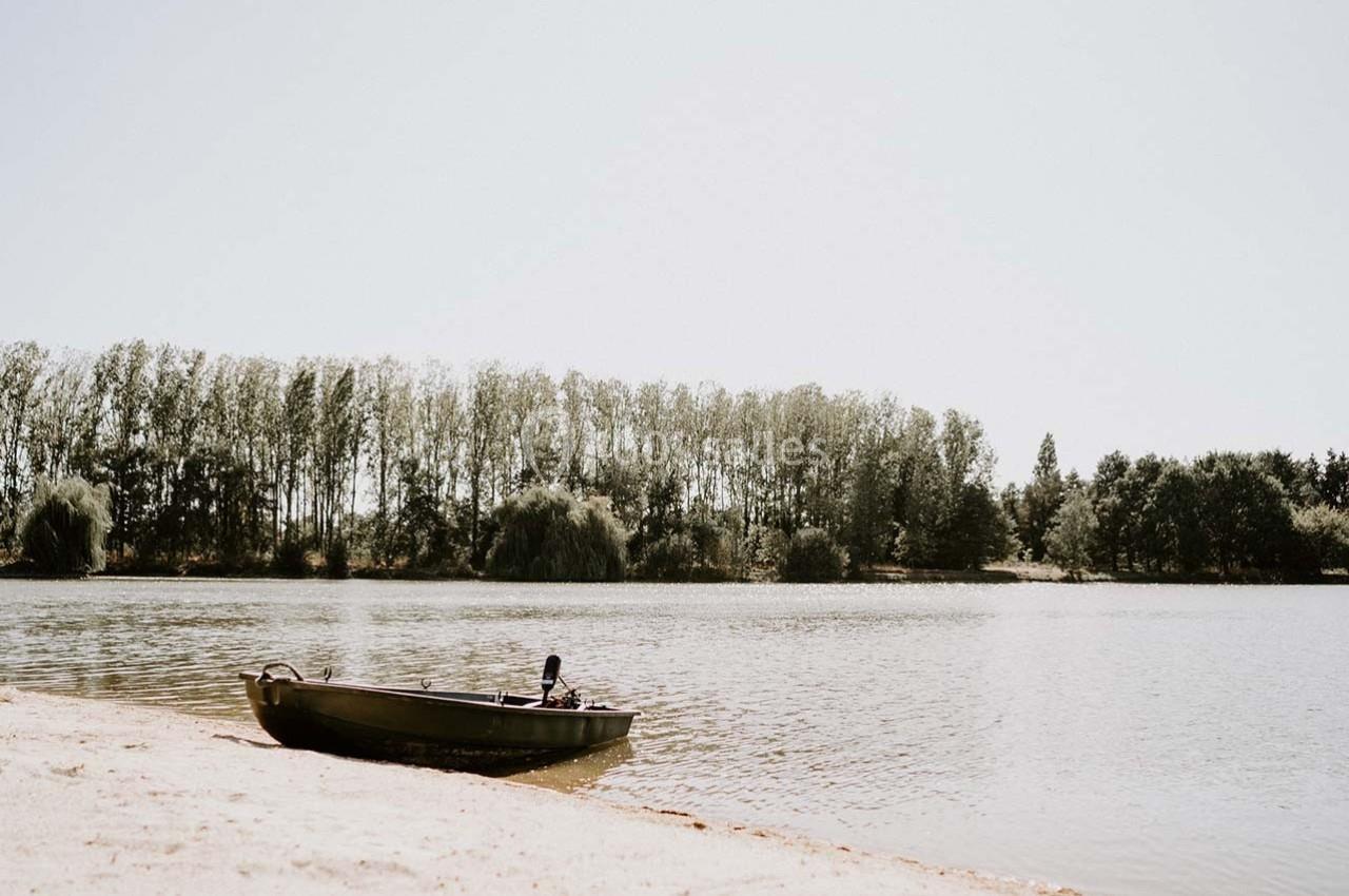 Une barque amarrée sur une plage au bord d'un lac, avec une rangée d'arbres en arrière-plan sous un ciel clair.