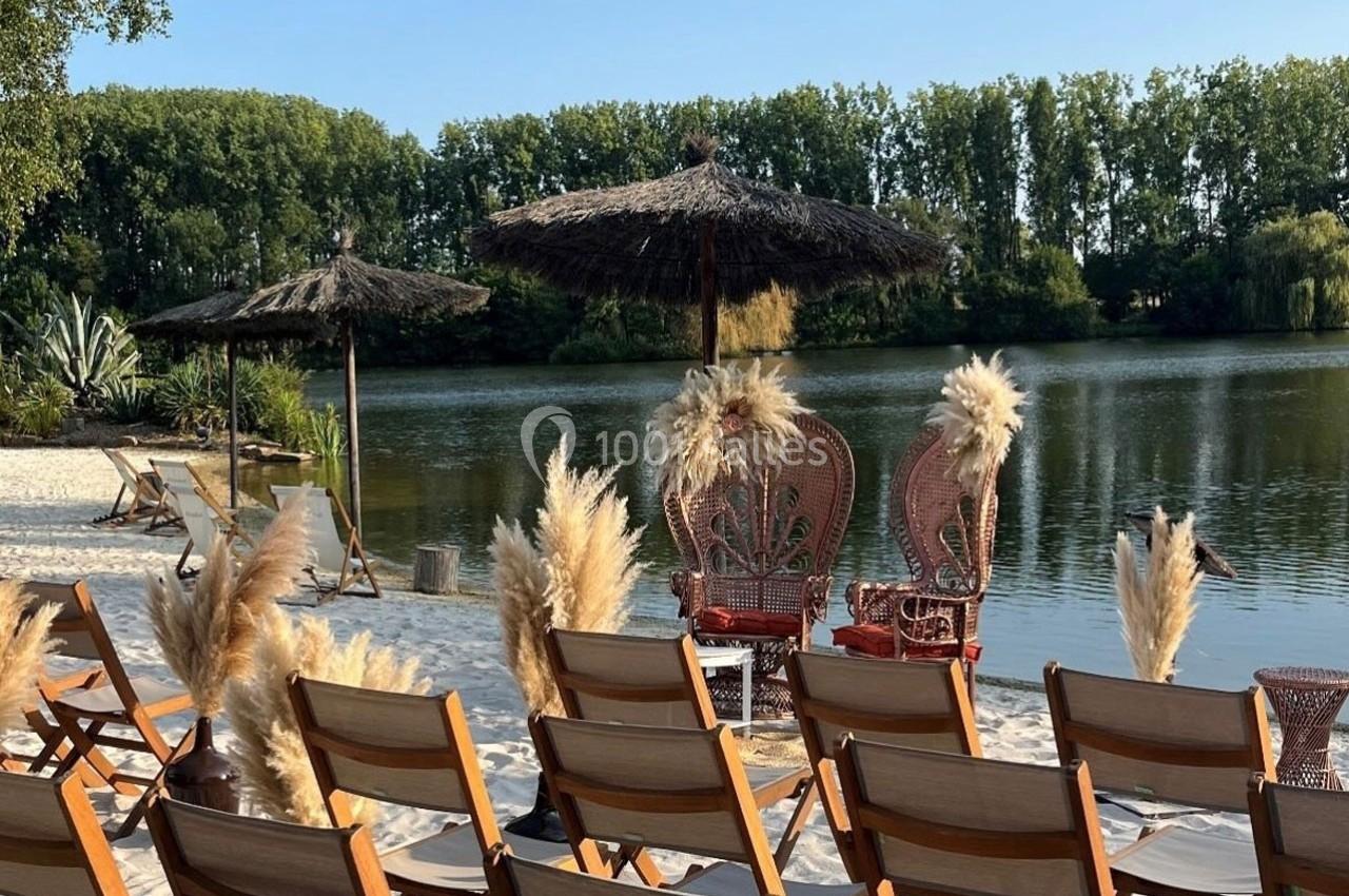 Chaises en bois alignées face à un lac, avec parasols en paille et décorations de fleurs séchées sur une plage de sable.