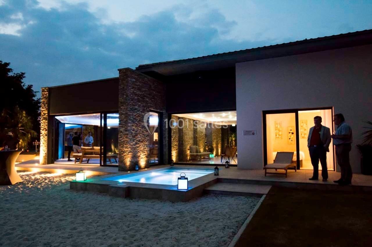 Maison moderne éclairée de nuit avec terrasse, piscine et invités debout à l'extérieur.