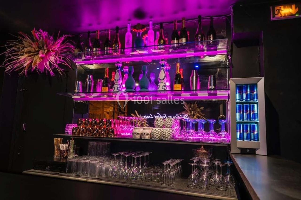 Bar avec étagères illuminées en rose, contenant bouteilles d'alcool, verres, et un réfrigérateur à boissons énergétiques.