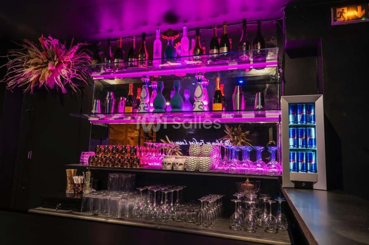 Étagères éclairées avec bouteilles d'alcool, verres et décorations dans un bar à l'ambiance lumineuse violette.