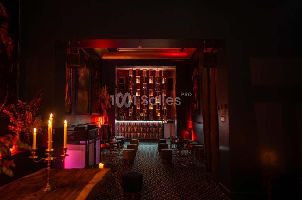 Salle sombre et élégante avec éclairage tamisé, bar illuminé en arrière-plan et chandeliers sur une table au premier plan.