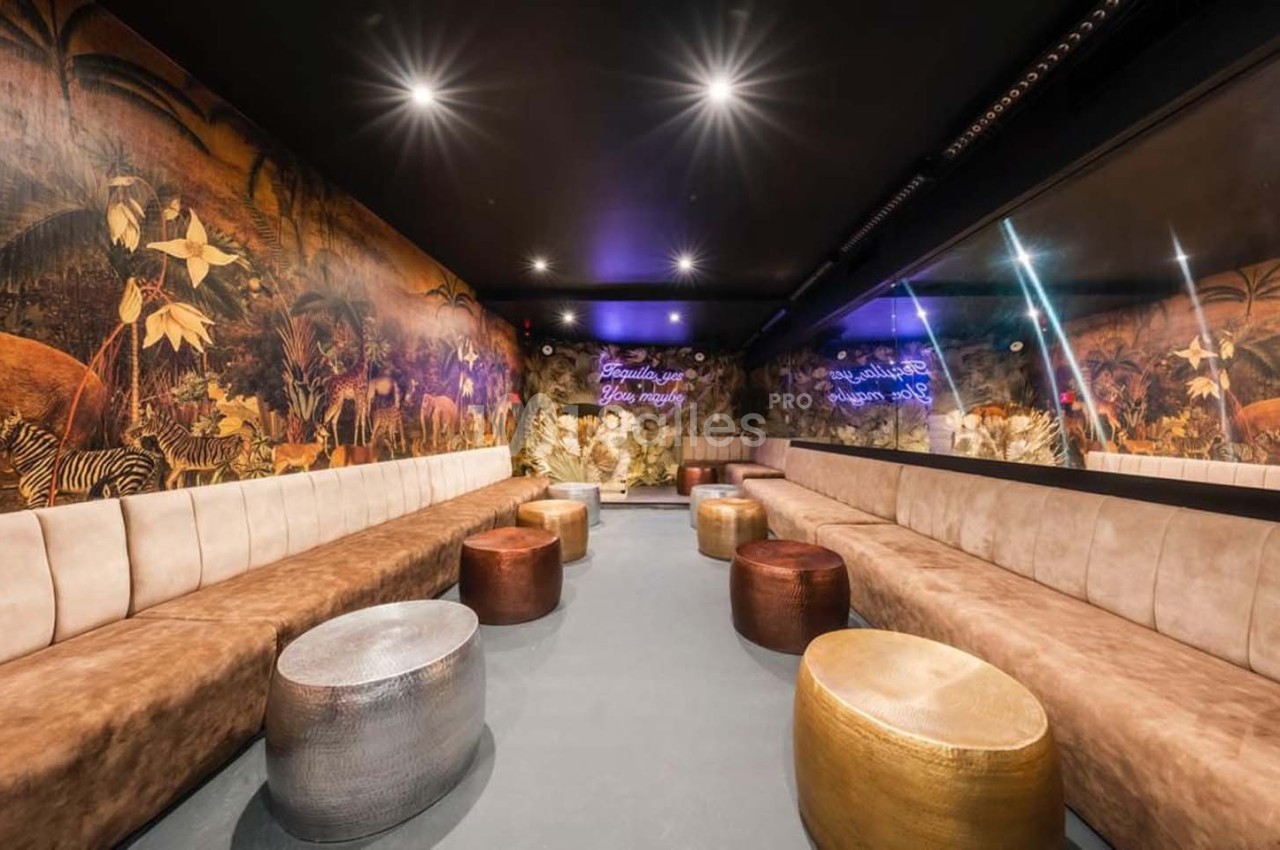 Salle lounge avec banquettes beige, tables basses métalliques et mur décoré de motifs tropicaux sous un éclairage tamisé.