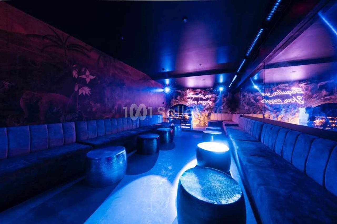 Salle lounge sombre avec banquettes, tables basses et éclairage bleu, décor mural tropical et néons lumineux.