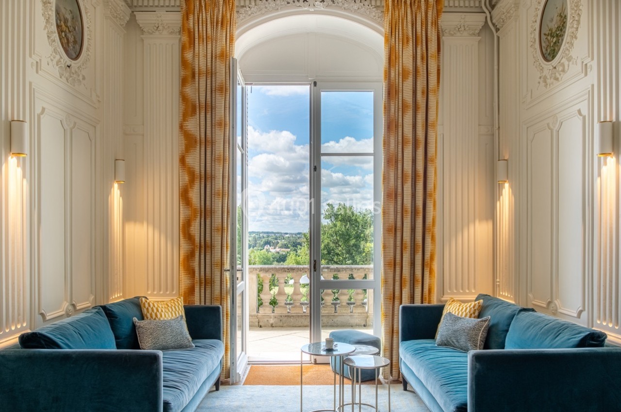 Salon lumineux avec deux canapés bleus, tables basses et grande baie vitrée donnant sur un balcon avec vue sur la nature.
