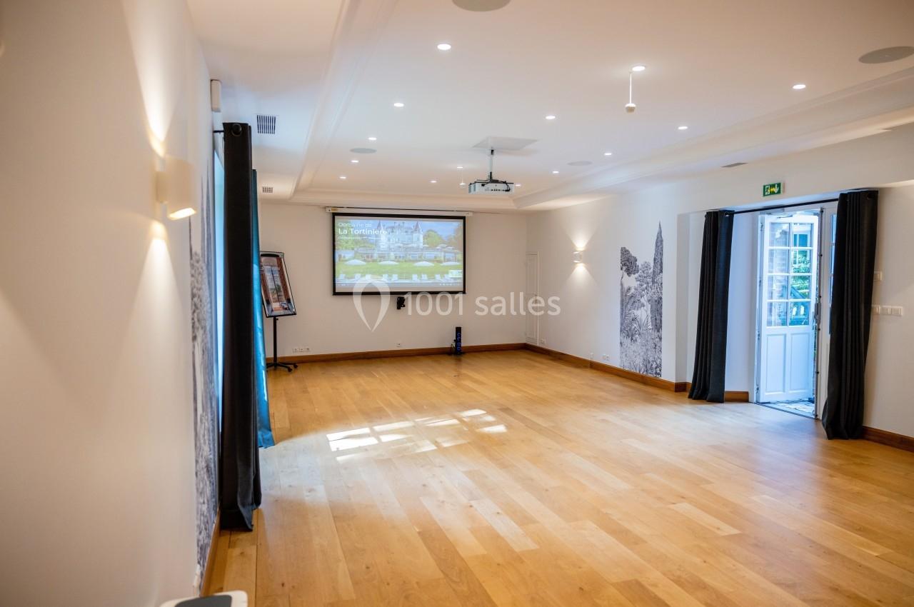 Salle lumineuse avec parquet en bois, écran de projection, rideaux sombres et porte ouverte donnant sur l'extérieur.