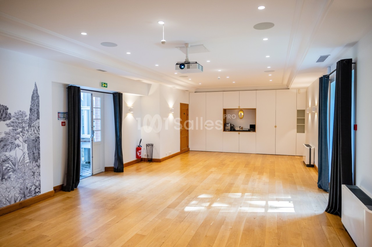 Salle lumineuse avec parquet en bois, murs blancs, rideaux sombres, projecteur au plafond et rangements intégrés.