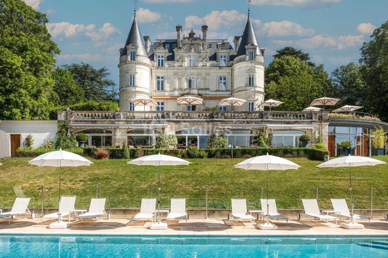 Façade d'un château entouré de verdure avec une piscine, des transats et des parasols au premier plan.