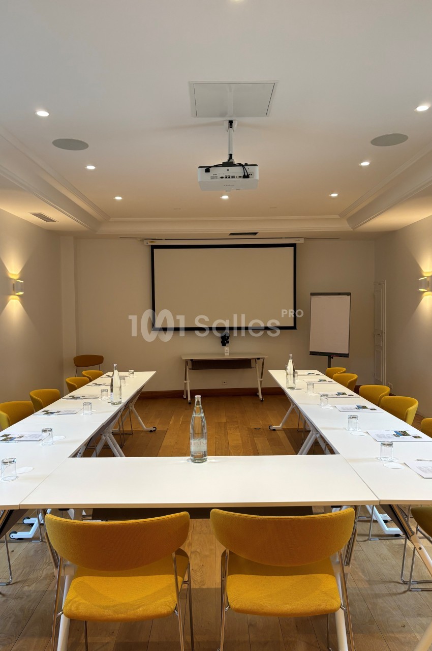 Salle de réunion lumineuse avec tables disposées en U, chaises jaunes, écran de projection et tableau blanc.