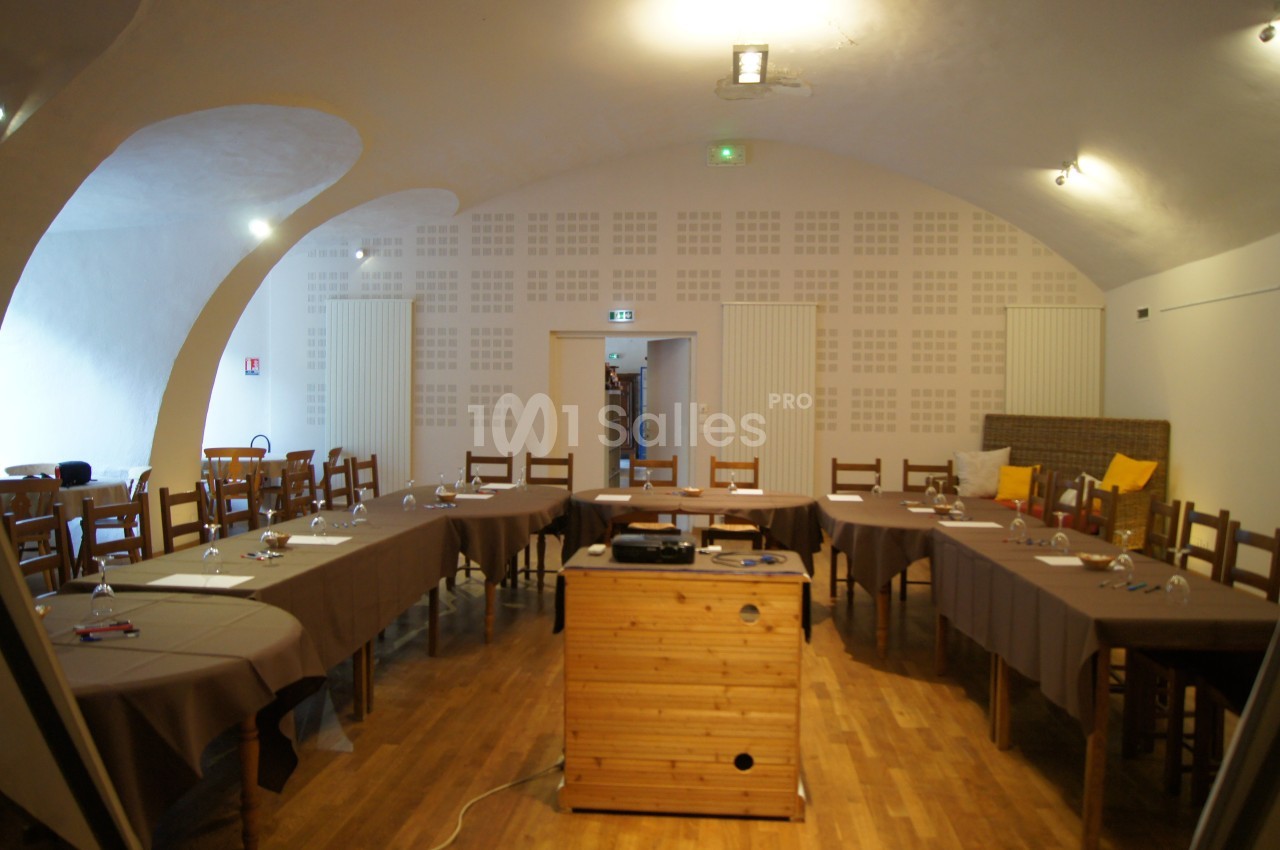 Salle de réunion avec tables disposées en U, chaises en bois, projecteur sur un meuble et éclairage au plafond.