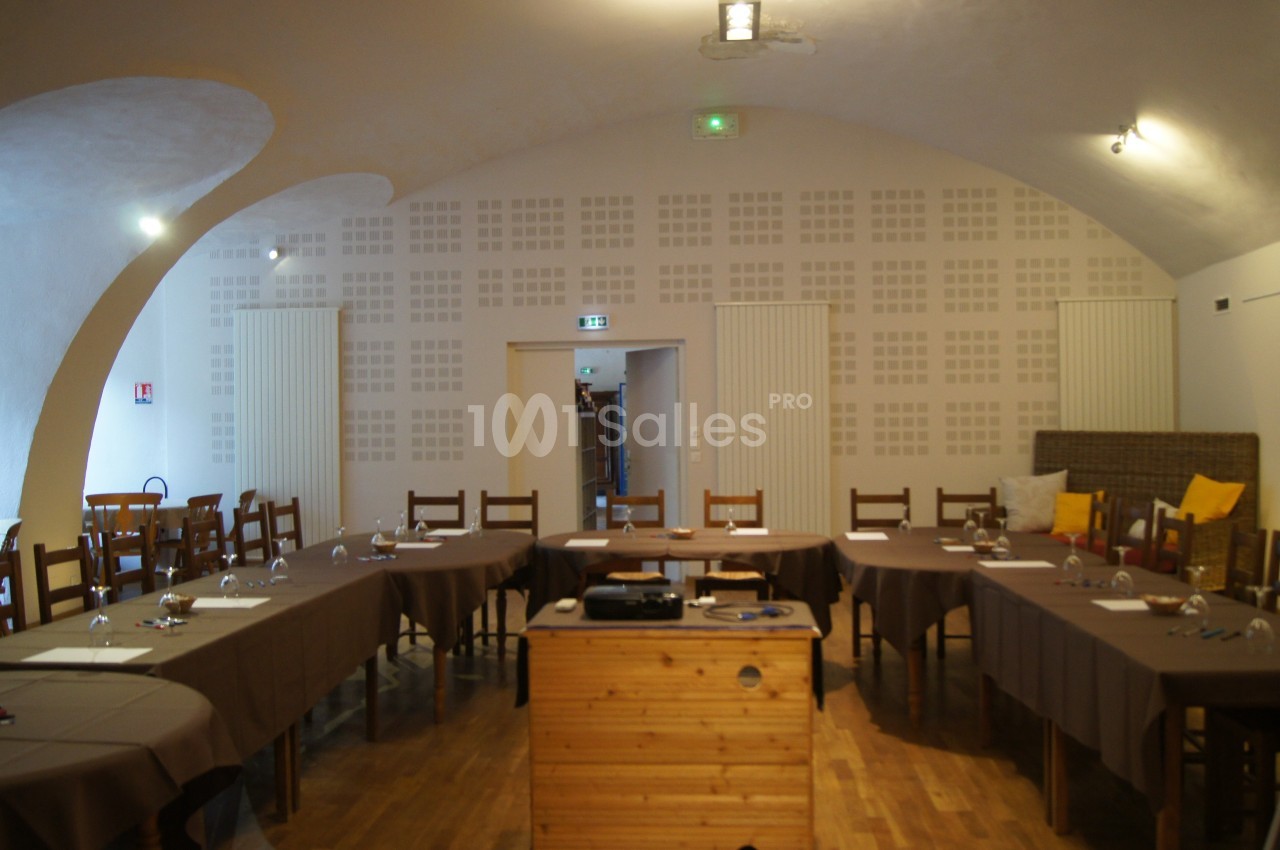 Salle de réunion avec tables disposées en U, chaises en bois, nappes brunes et projecteur sur un meuble en bois.