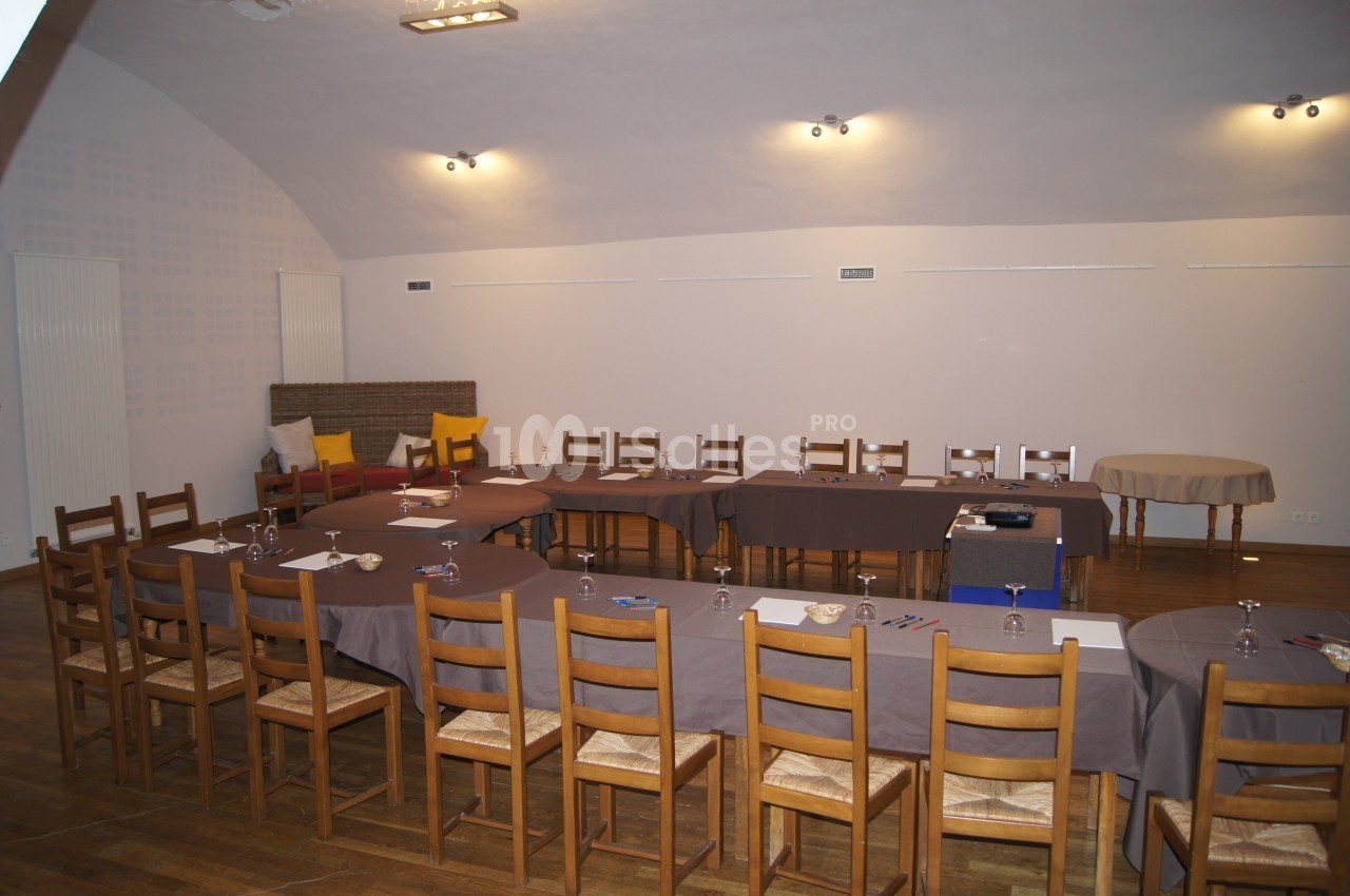 Salle de réunion aménagée en U avec des tables recouvertes de nappes grises, chaises en bois et matériel de travail.