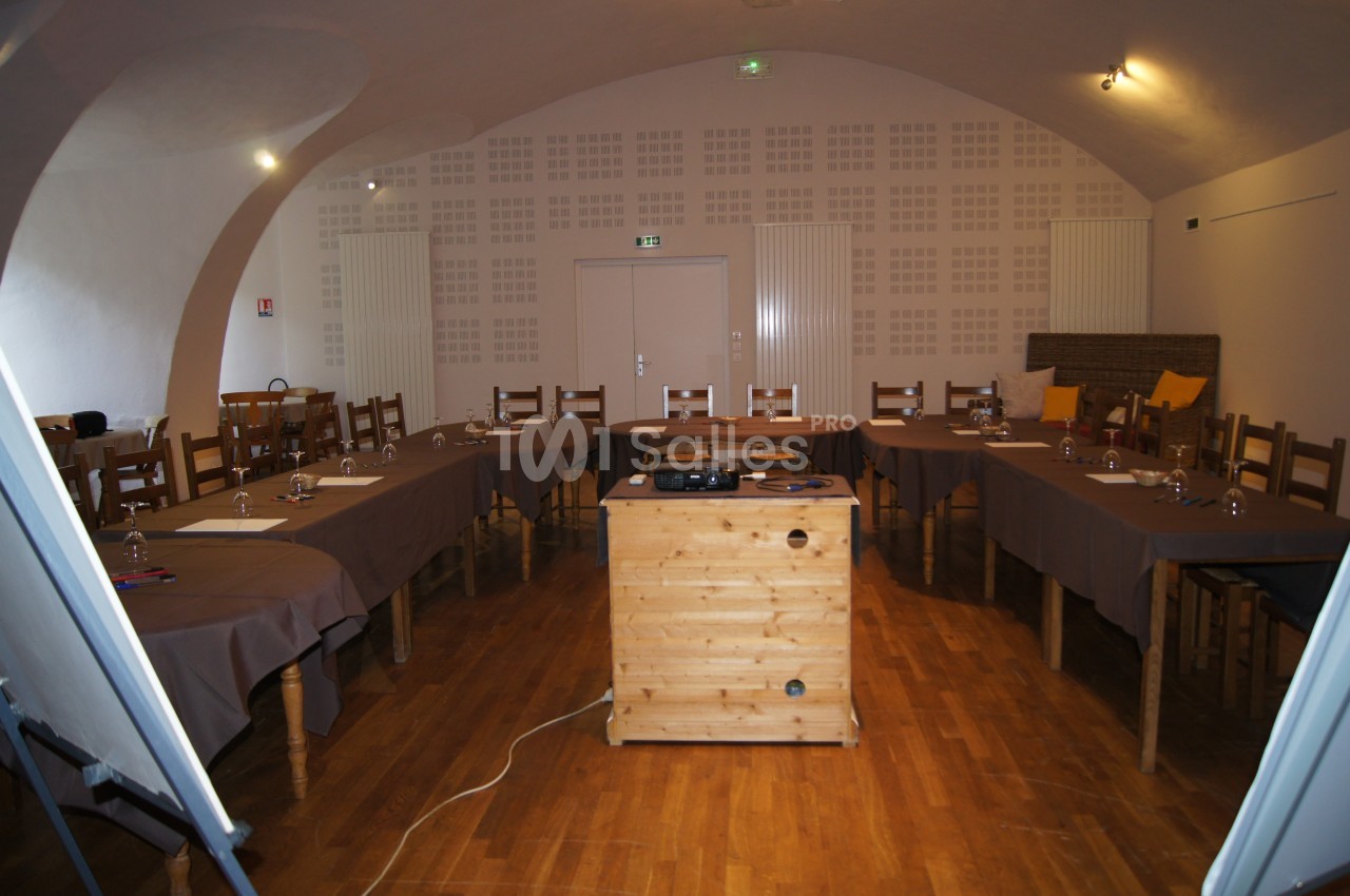 Salle de réunion avec tables disposées en U, chaises en bois, projecteur sur un meuble central et éclairage tamisé.