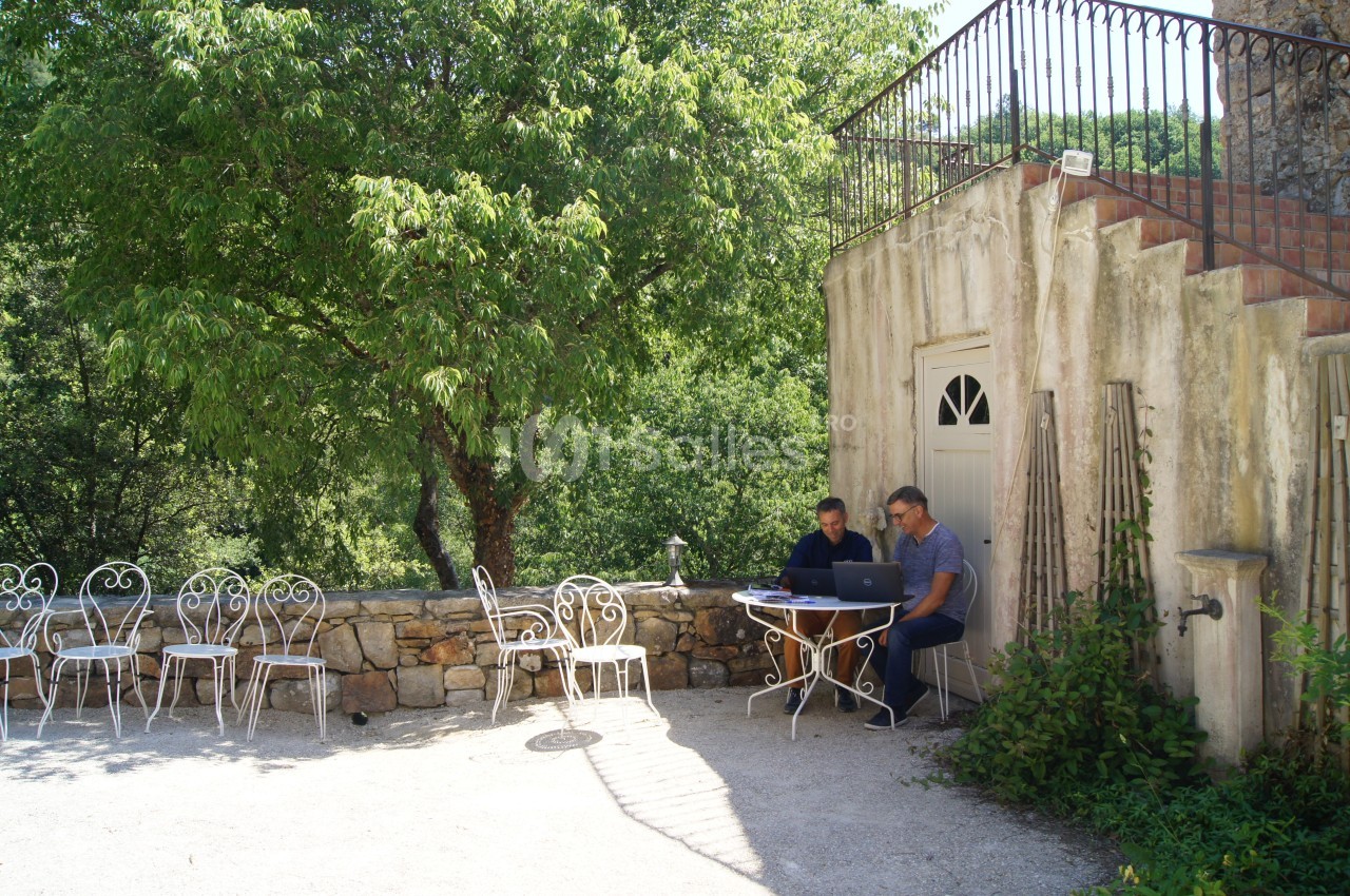 Deux personnes travaillent sur un ordinateur portable à une table en extérieur, près d'un bâtiment en pierre et d'arbres.