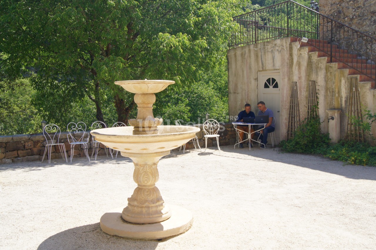 Fontaine en pierre dans une cour ensoleillée, avec des tables et des chaises en fer forgé près d'un bâtiment ancien.