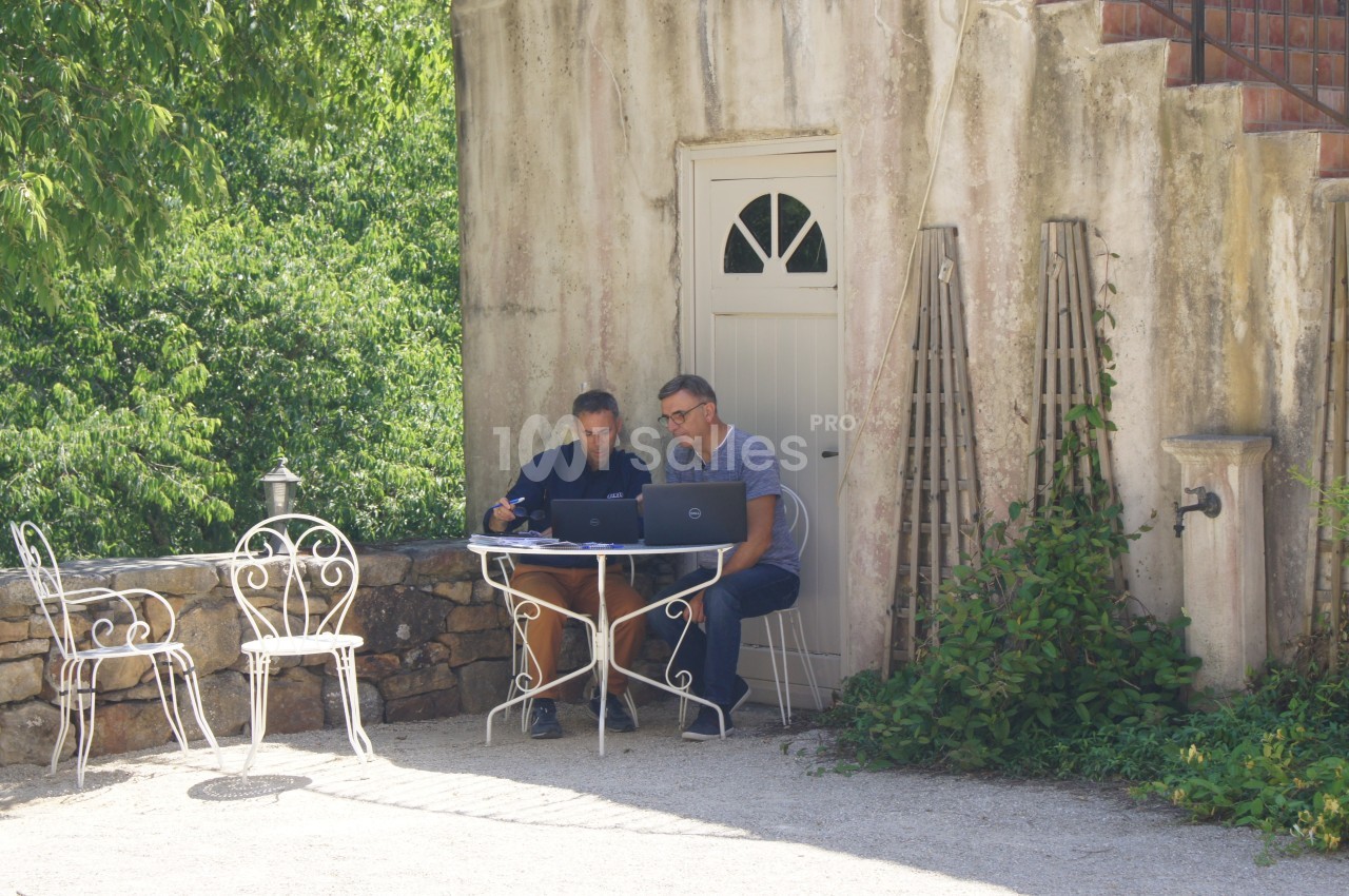 Deux hommes travaillent sur un ordinateur portable à une table en fer forgé, devant une maison en pierre entourée de verdure.