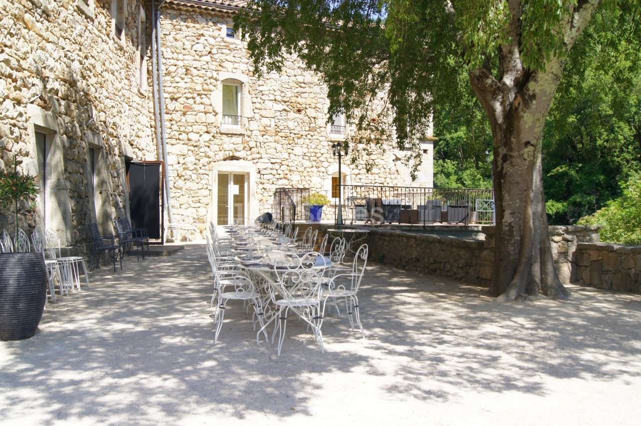 Terrasse ensoleillée avec tables et chaises en métal blanc devant un bâtiment en pierre et un grand arbre.