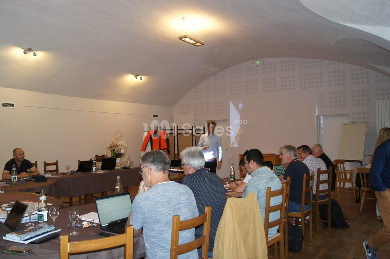 Un groupe de personnes assises autour de tables en réunion dans une salle éclairée avec un présentateur debout.