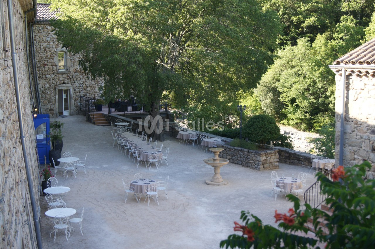 Cour extérieure avec tables et chaises en fer forgé, fontaine centrale, arbres et bâtiments en pierre environnants.