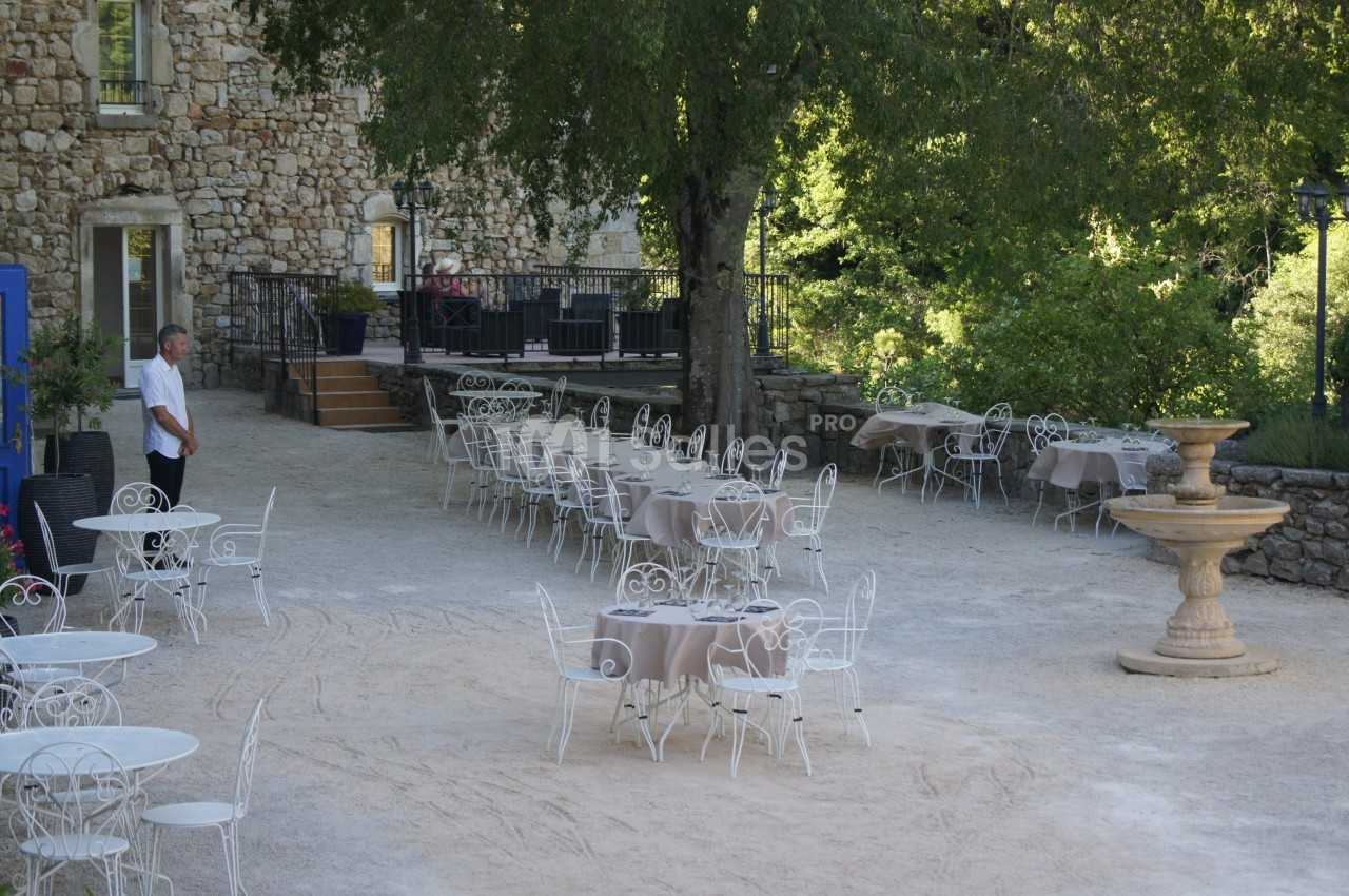 Terrasse extérieure avec tables et chaises en fer forgé, entourée de verdure et d'un bâtiment en pierre.