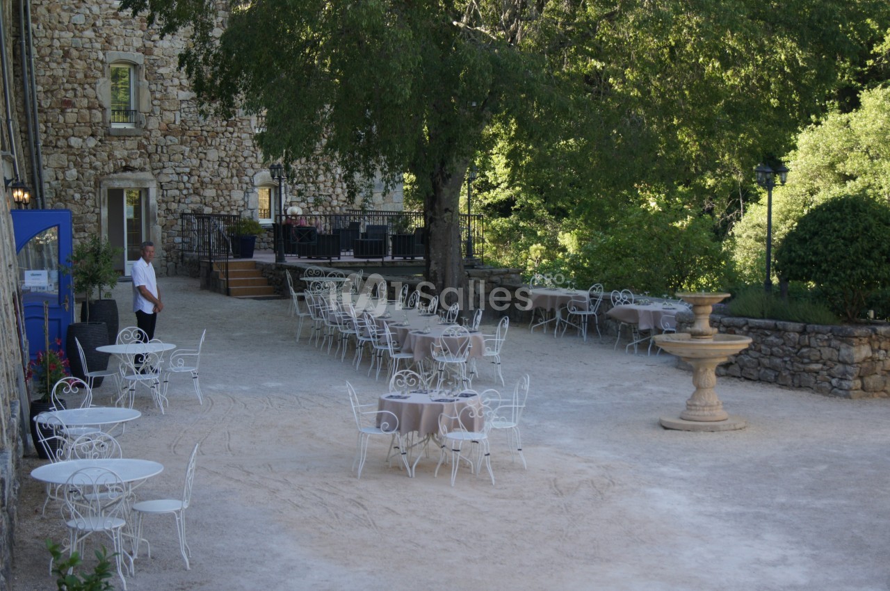 Cour extérieure avec tables et chaises en fer forgé, fontaine centrale et bâtiment en pierre sous des arbres.