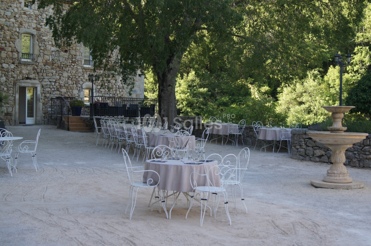 Terrasse extérieure avec tables et chaises en fer forgé, entourée de verdure et d'un bâtiment en pierre.
