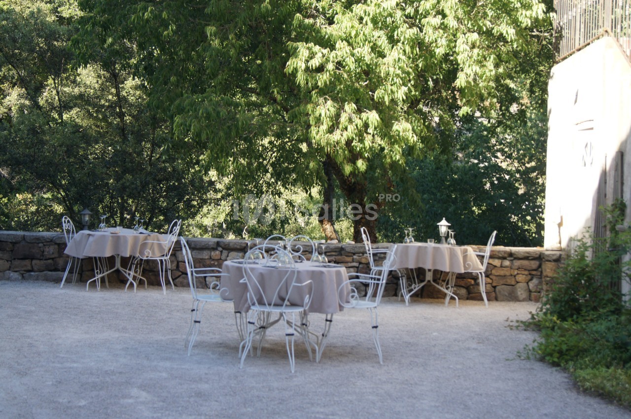 Tables en fer forgé avec nappes beiges disposées en extérieur sur une terrasse ombragée près d'un mur en pierre.