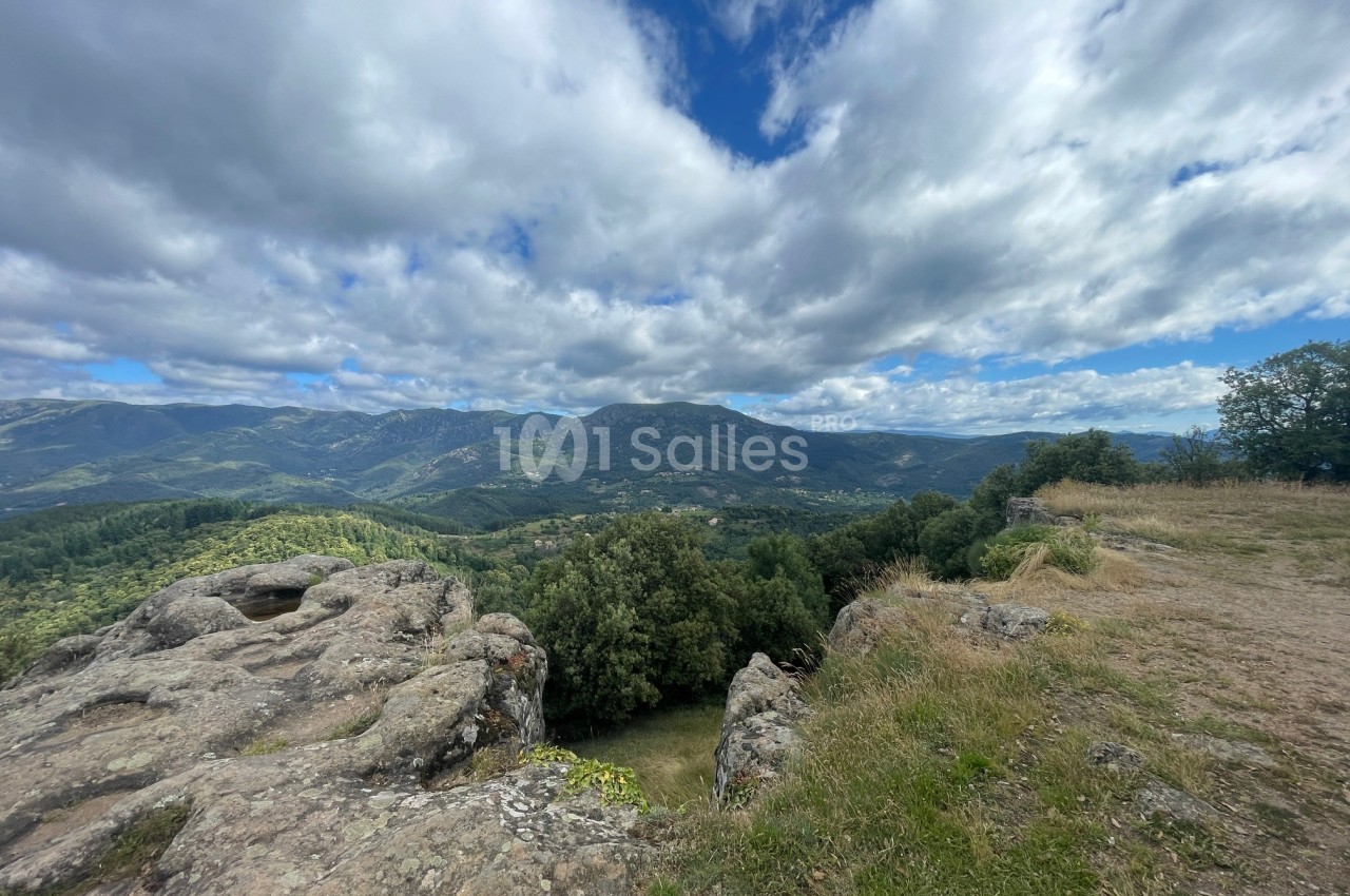 Paysage de montagne avec des rochers au premier plan, une vallée verdoyante et un ciel partiellement nuageux.