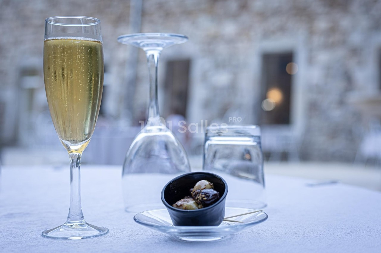 Verre de champagne, verrines et verrerie renversée sur une table blanche en extérieur avec un mur en pierre en arrière-plan.
