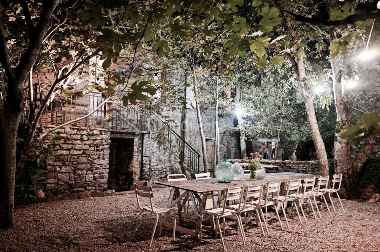Grande table en bois entourée de chaises dans une cour extérieure éclairée, entourée d'arbres et de murs en pierre.