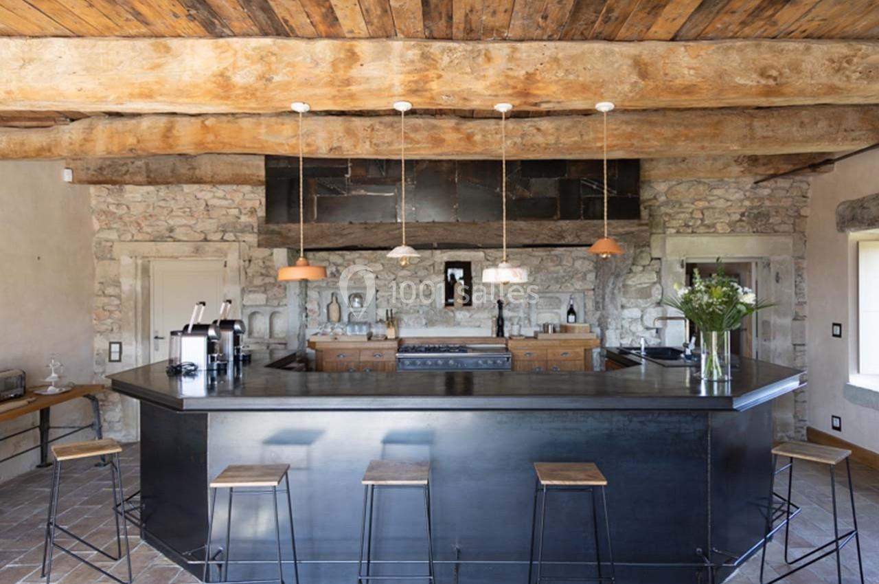 Cuisine rustique avec murs en pierre, poutres en bois apparentes, îlot central noir et tabourets en métal.
