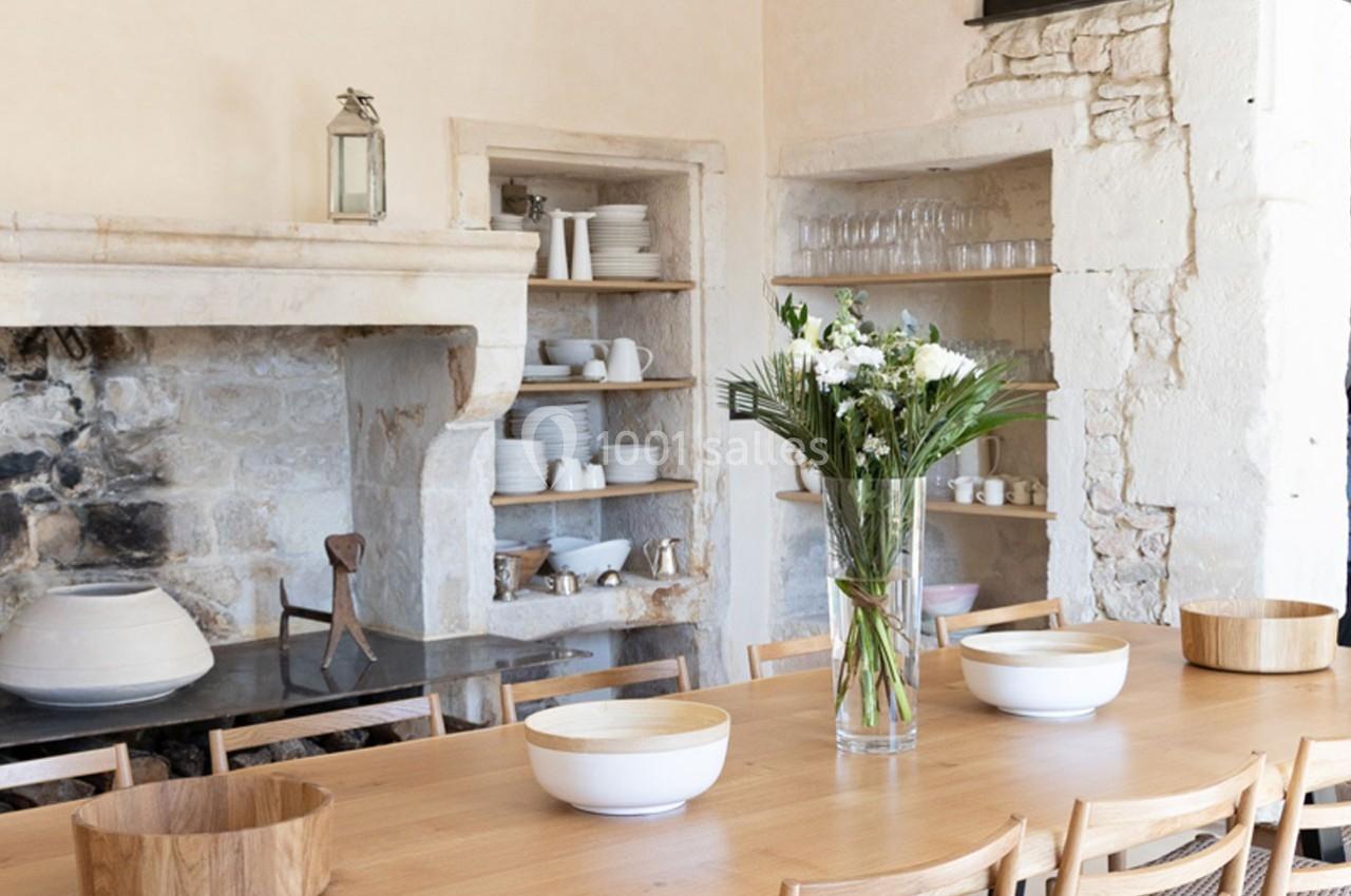 Grande table en bois entourée de chaises, décorée avec un vase de fleurs, dans une cuisine rustique avec étagères ouvertes.