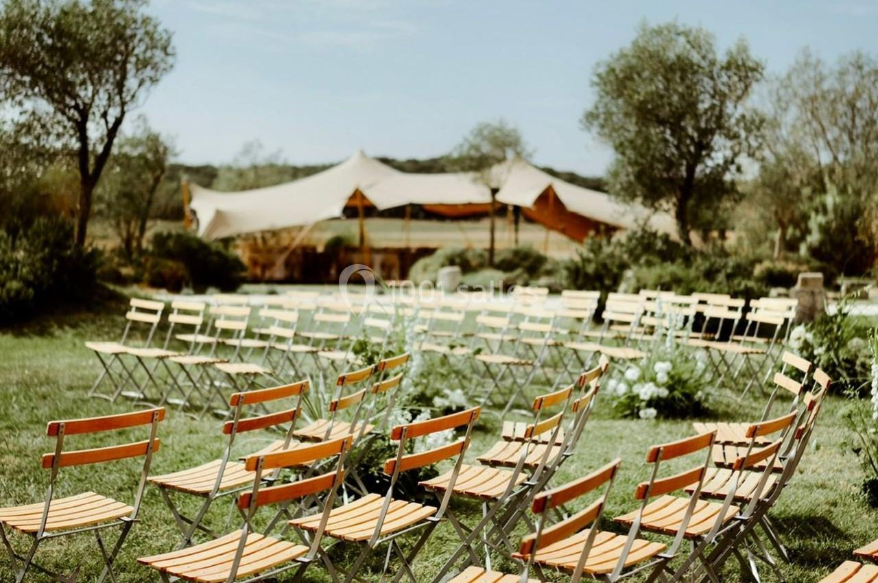Chaises pliantes disposées en rangées sur une pelouse, avec une tente beige et des arbres en arrière-plan.