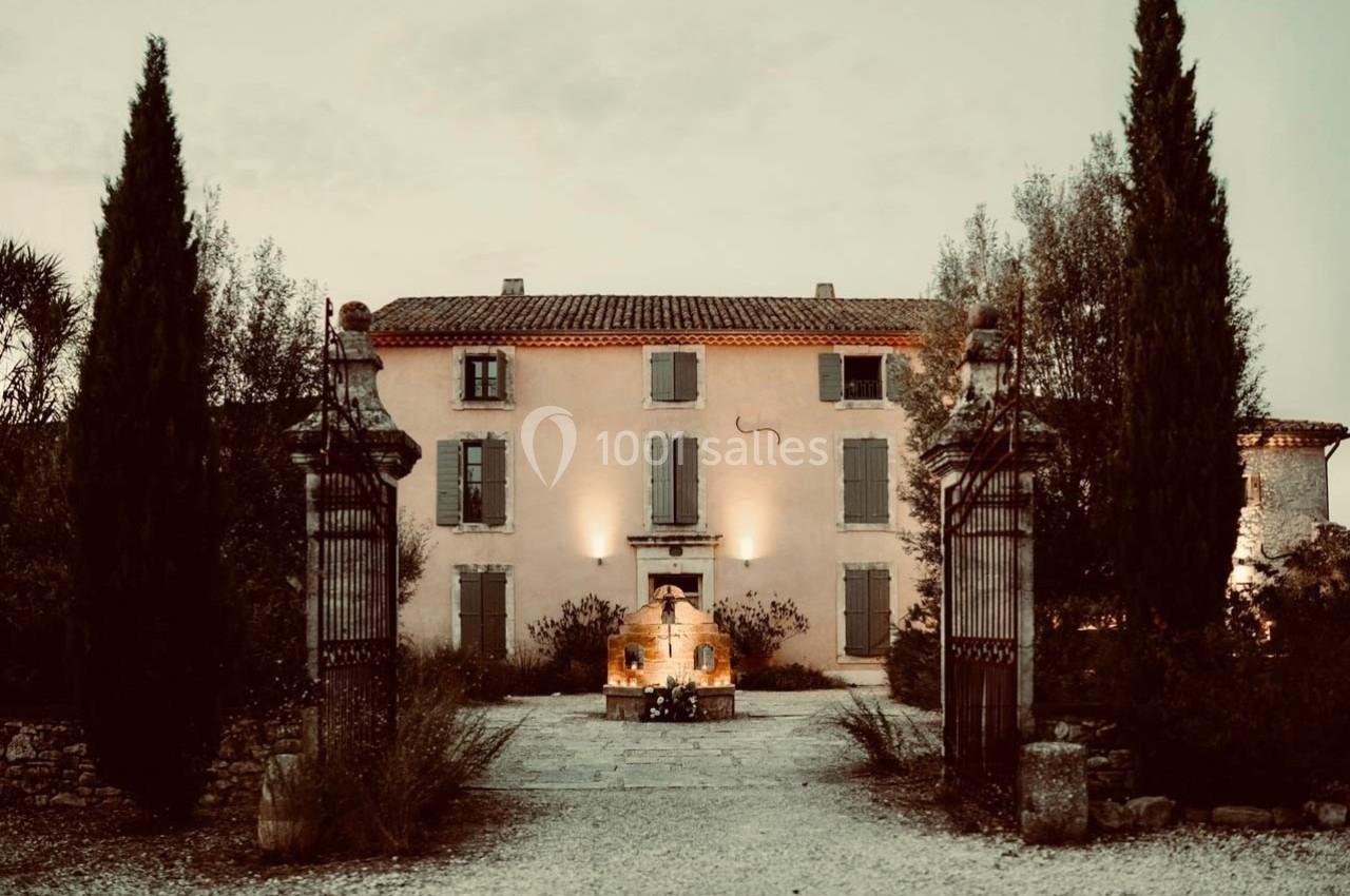 Façade d'une maison de campagne en pierre, entourée de cyprès et d'un portail en fer forgé, au crépuscule.
