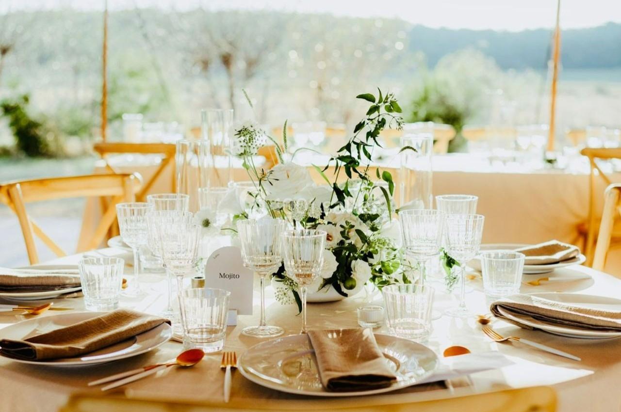 Table élégamment dressée avec vaisselle en verre, serviettes beige et centre de table floral dans un cadre extérieur…