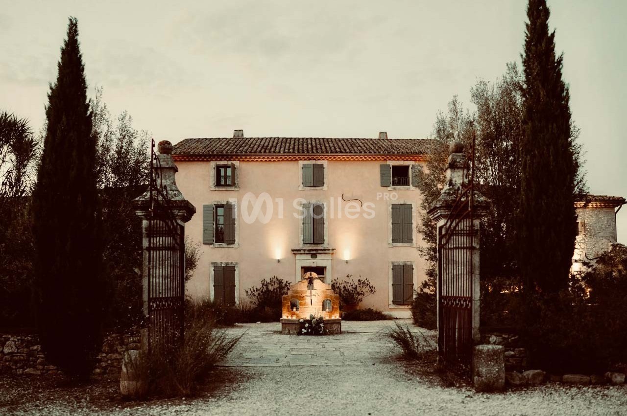 Façade d'une maison de campagne en pierre, entourée de cyprès et d'un portail en fer forgé, au crépuscule.