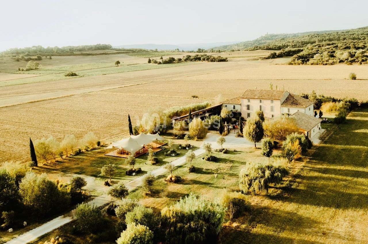 Vue aérienne d'une grande maison entourée de champs et d'arbres dans un paysage rural ensoleillé.