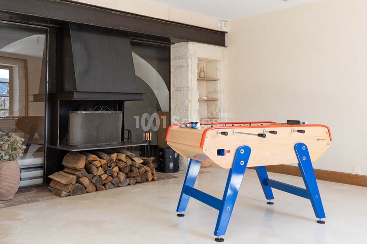 Baby-foot en bois avec pieds bleus dans une pièce lumineuse, près d'une cheminée moderne et d'un tas de bois.