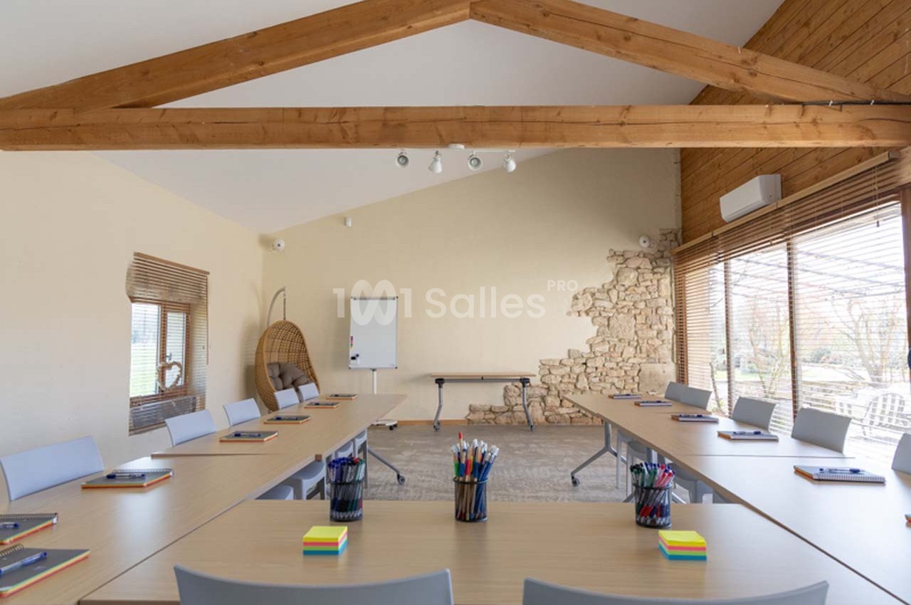 Salle de réunion lumineuse avec tables disposées en U, tableau blanc, chaises et grandes fenêtres donnant sur l'extérieur.