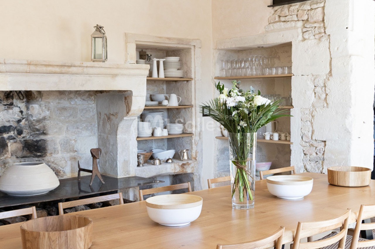 Grande table en bois entourée de chaises, décorée de fleurs, dans une cuisine rustique avec étagères et cheminée en pierre.