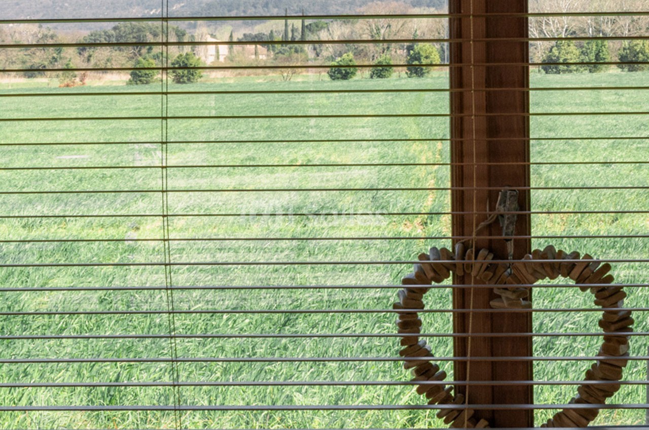 Vue d'un champ verdoyant à travers une fenêtre avec des stores en bois et une décoration en forme de cœur suspendue.