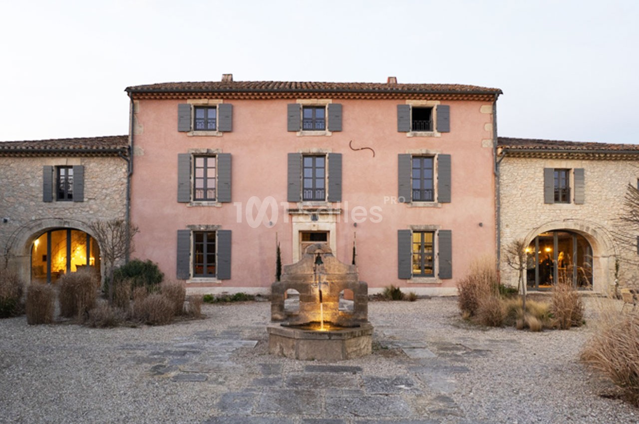Façade d'une grande maison en pierre avec volets gris, une fontaine centrale et une cour gravillonnée.