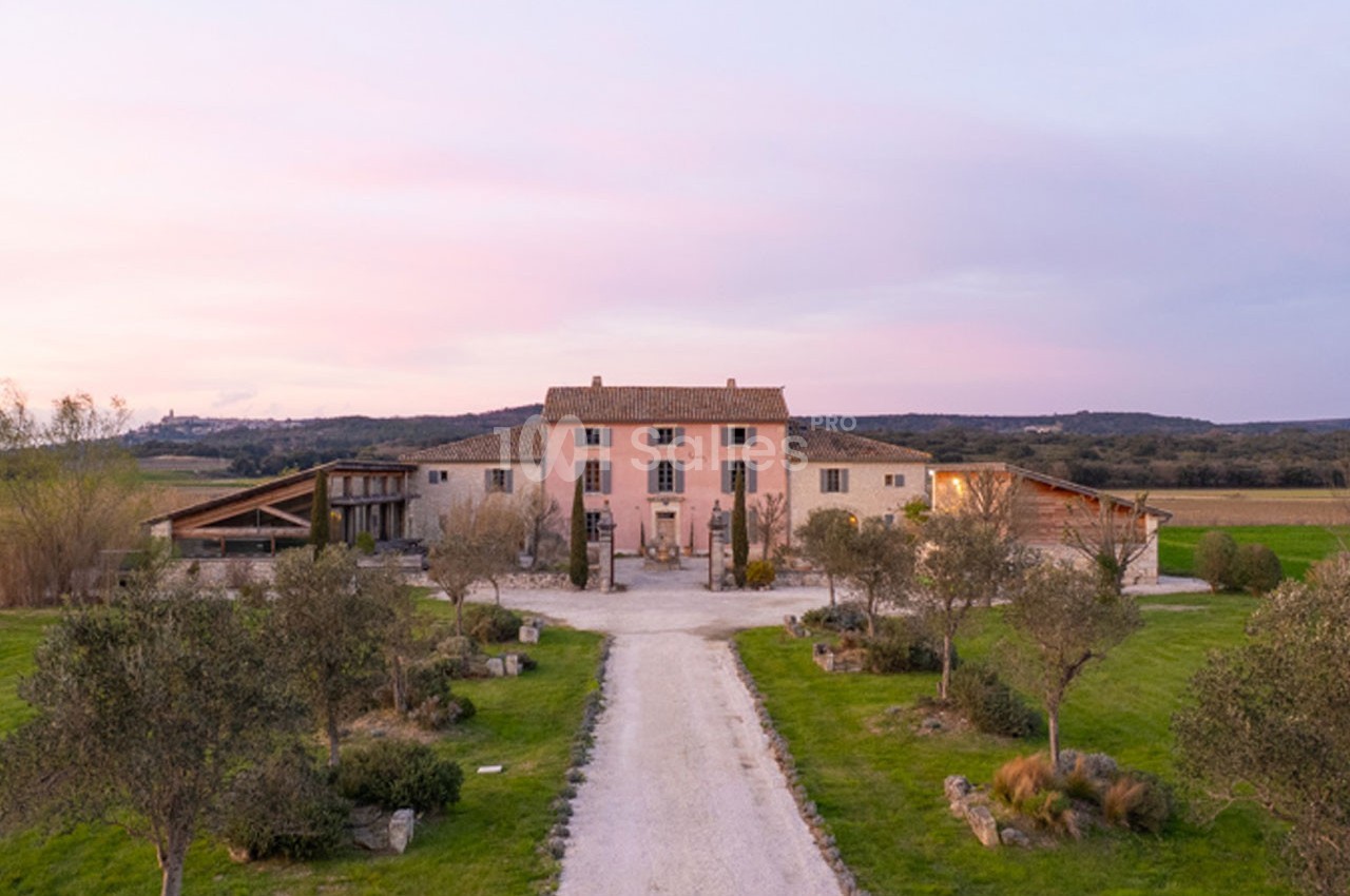 Allée bordée d'oliviers menant à une grande maison en pierre entourée de champs sous un ciel au coucher du soleil.