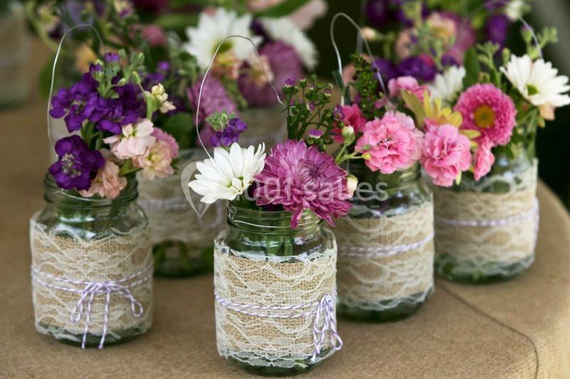 Des pots en verre décorés de dentelle contenant des bouquets de fleurs colorées sur une table en tissu brun.