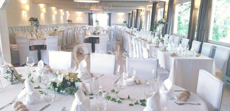 Salle de réception lumineuse avec tables décorées de nappes blanches, serviettes pliées et arrangements floraux.