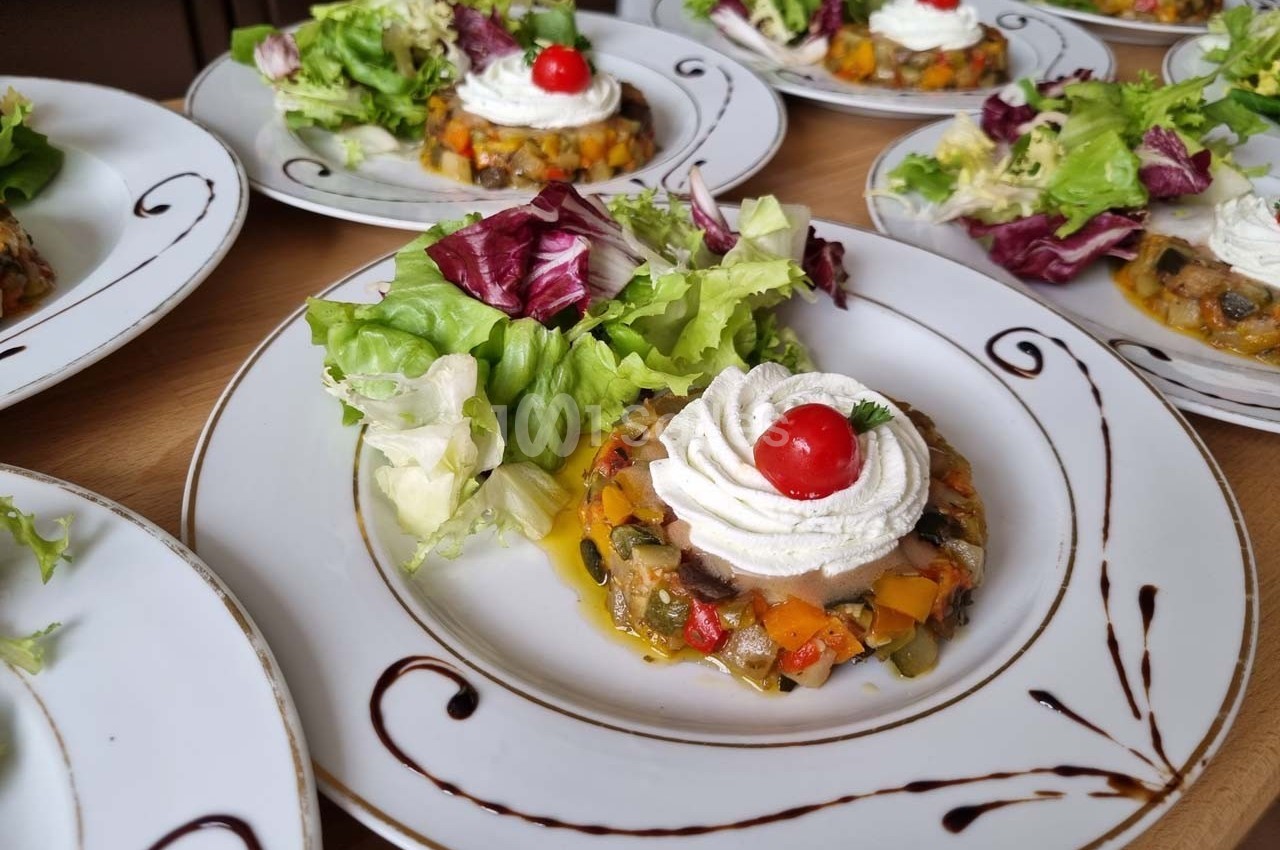 Assiette contenant une ratatouille dressée en cercle, surmontée de chantilly salée et d'une tomate cerise, accompagnée de…