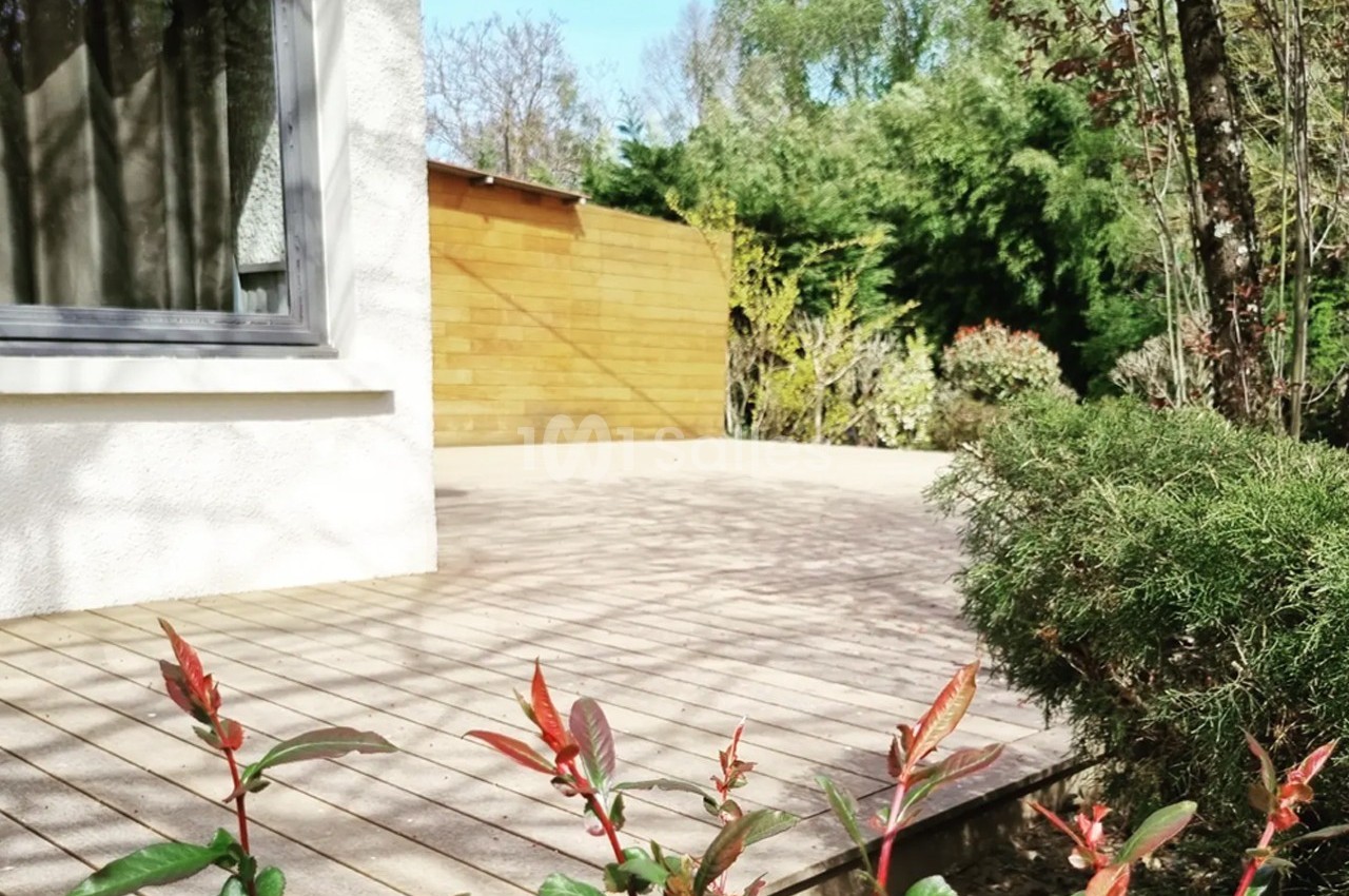 Terrasse en bois attenante à une maison blanche, entourée de végétation et baignée de lumière naturelle.