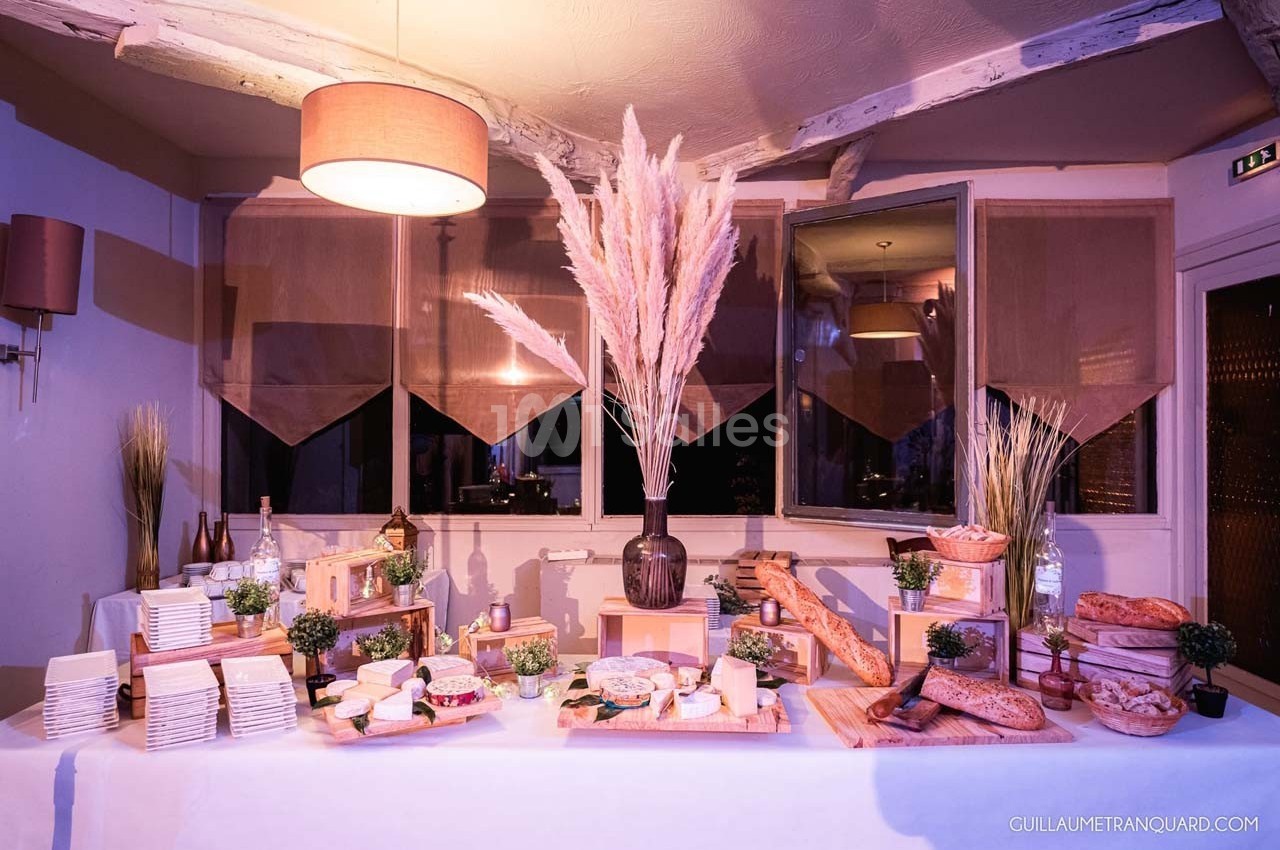 Table garnie de fromages, pains et décorations végétales dans une salle éclairée par une lumière chaleureuse.