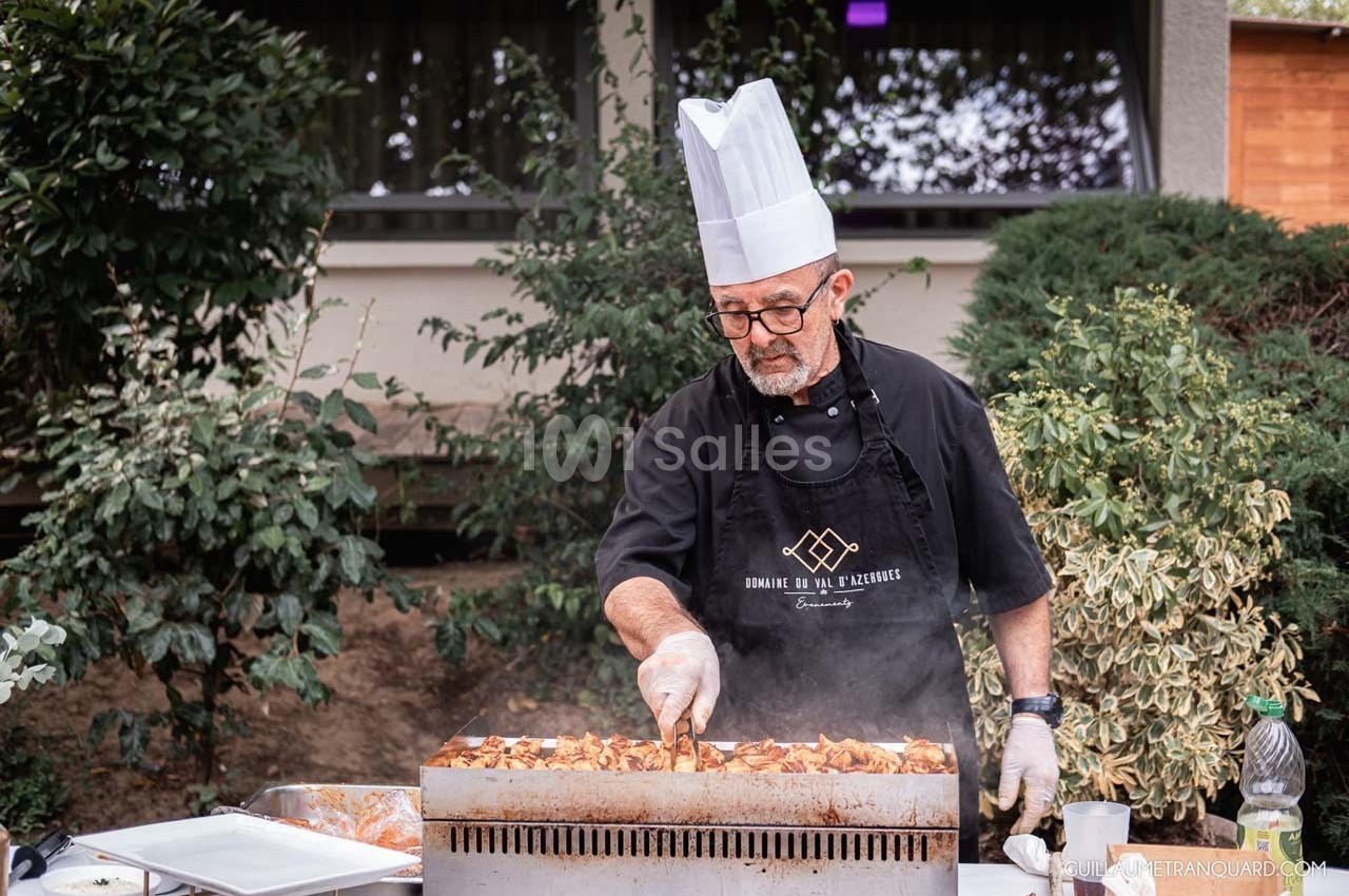 Un chef en tablier et toque cuisine des légumes sur une grande plaque chauffante en extérieur.