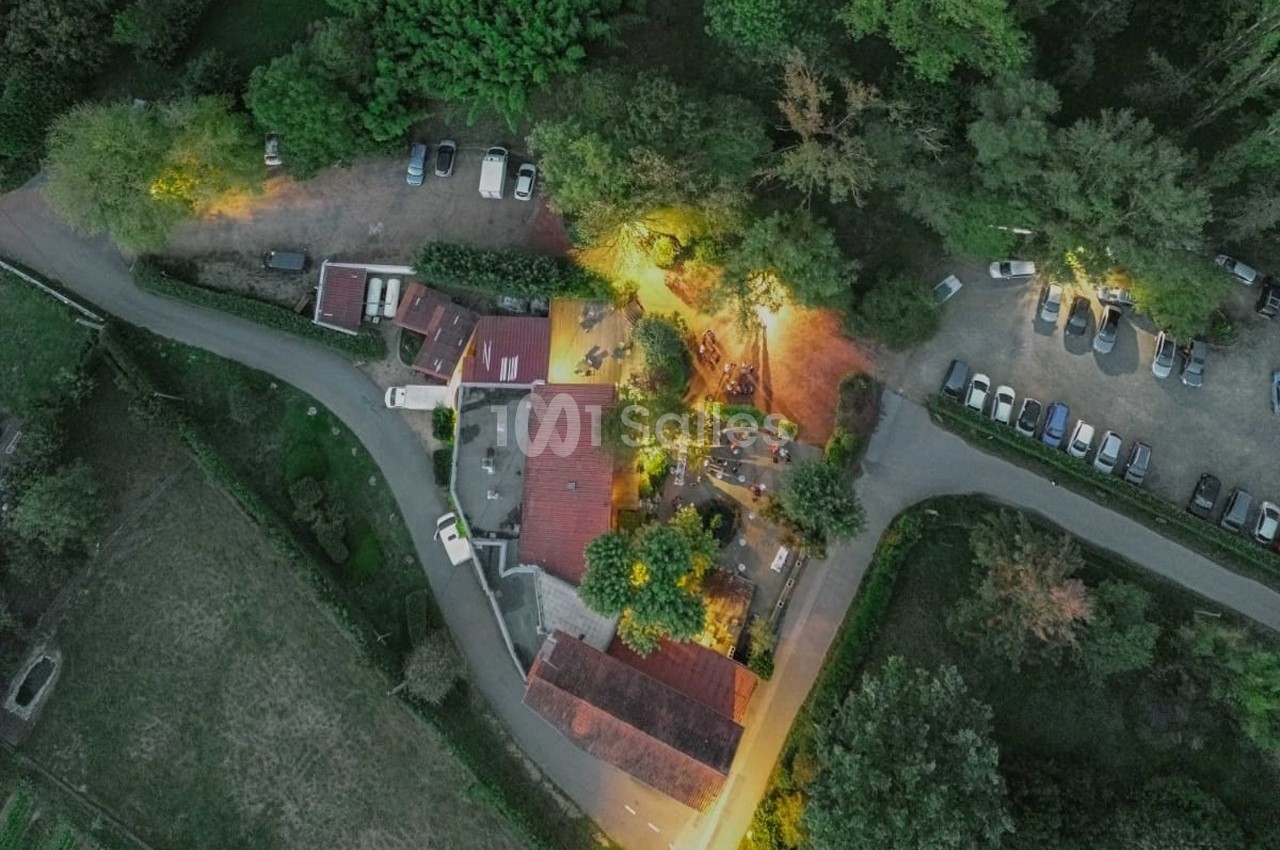 Vue aérienne d'un bâtiment entouré de végétation, avec des voitures garées et des chemins éclairés en soirée.